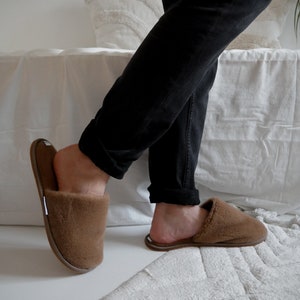 May include: A pair of brown fuzzy slippers with a closed toe and an open heel. The slippers are worn by a person wearing black pants.
