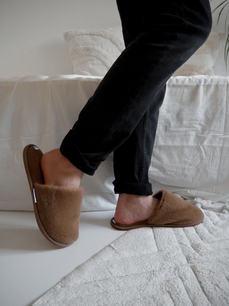 May include: A pair of brown fuzzy slippers with a closed toe and an open heel. The slippers are worn by a person wearing black pants.