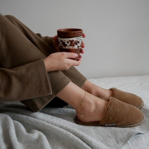 May include: A person wearing brown pajamas and fuzzy slippers sits on a white bed, holding a brown ceramic mug with white designs.