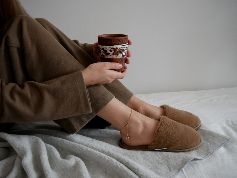 May include: A person wearing brown pajamas and fuzzy slippers sits on a white bed, holding a brown ceramic mug with white designs.