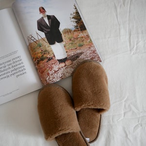 May include: A pair of brown fuzzy slippers with a closed toe and an open back. The slippers are on a white surface.