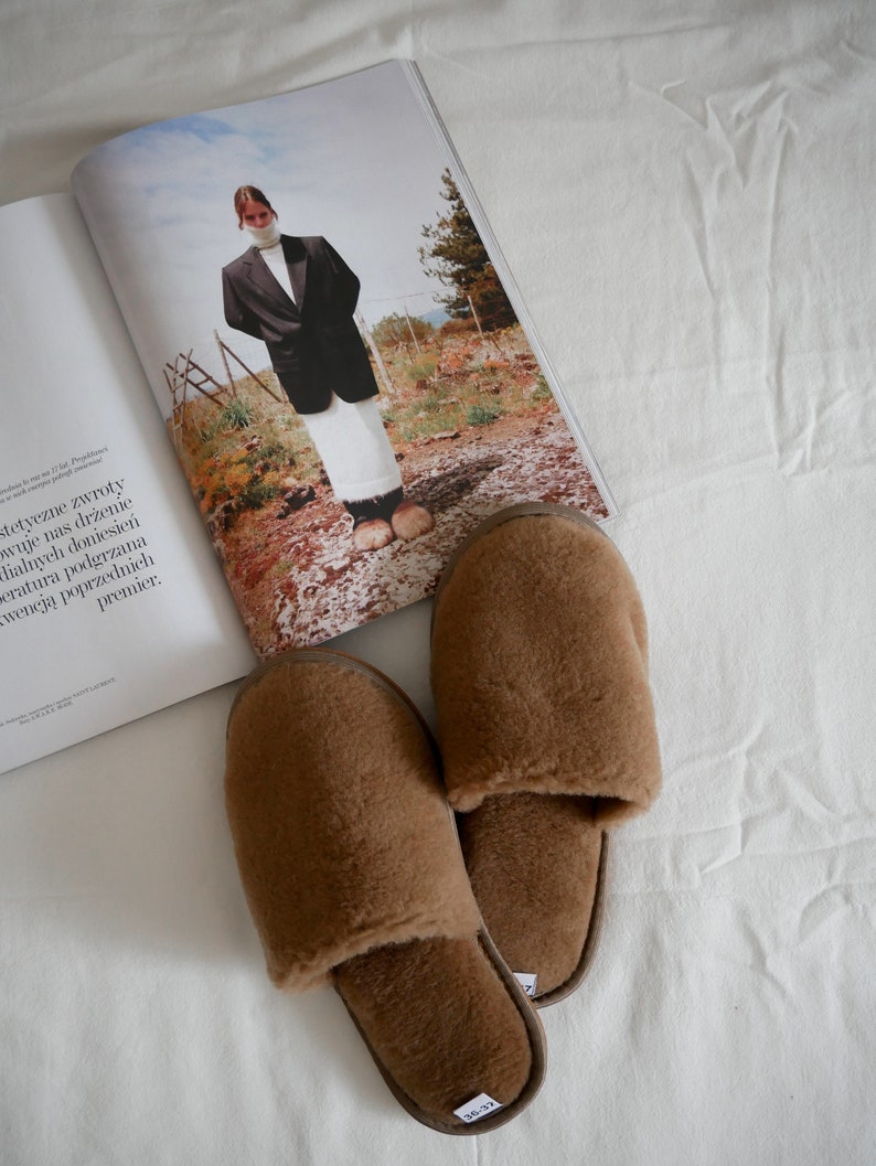 May include: A pair of brown fuzzy slippers with a closed toe and an open back. The slippers are on a white surface.