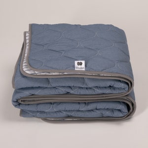 Op de afbeelding: Een gevouwen blauwe quilt met een grijze rand en een wit label met de tekst "Woollen" in zwarte letters.