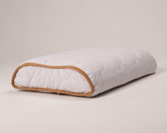 May include: A white pillow with a quilted pattern and a brown trim. The pillow is shaped like a crescent moon.