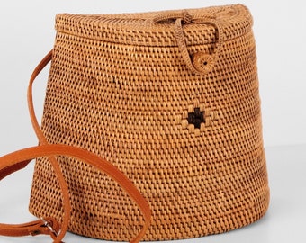 May include: A woven rattan crossbody bag in a natural tan color. The bag has a rounded top, a leather strap, and a decorative dark brown cross-shaped design on the front. The bag is designed with a clasp closure.