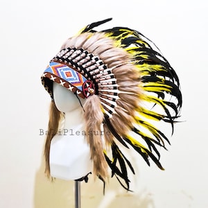 May include: A Native American headdress with a white, brown, yellow, and black feather design. The headdress has a beaded band with a geometric pattern in blue, orange, and white. The text "BaliPleasure" is visible.