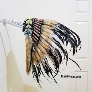 Könnte beinhalten: Ein indianischer Kopfschmuck mit einem schwarz, gelb und blau bestickten Band. Der Kopfschmuck hat braune, schwarze und weiße Federn und einen Fellakzent. Der Text "BaliPleasure" ist auf der rechten Seite des Bildes zu sehen.