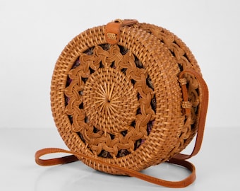 May include: A round, woven rattan handbag with a brown leather strap. The bag features a decorative, openwork design around the edges and a woven center. The bag is a natural tan color and has a leather closure.