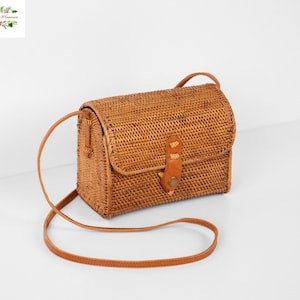 May include: A rectangular woven rattan crossbody bag in a natural brown tone. The bag features a structured flap closure secured with a tan leather strap and a metal snap button. It includes a thin, matching tan leather shoulder strap.