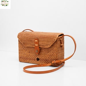 May include: A light brown rectangular crossbody bag made of woven rattan. The bag features a flap closure with a leather strap and buckle.  It has a short, adjustable leather shoulder strap. The bag is displayed on a white background.