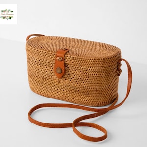 May include: An oval-shaped crossbody bag crafted from woven light brown rattan material. The bag features a structured lid, a tan leather strap closure with a single button, and a matching thin leather shoulder strap attached to the sides.