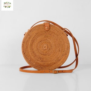 May include: A round, woven rattan crossbody bag with a brown leather strap and buckle. The bag has a spiral design in the center and a leather flap closure. The bag is a light brown color.