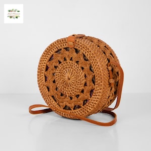 May include: A round, woven rattan handbag with a brown leather strap. The bag features a decorative, openwork design around the edges and a woven center. The bag is a natural tan color and has a leather closure.
