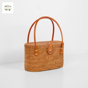 May include: Woven rattan handbag with a rectangular shape and rounded edges. The bag features a leather strap and a leather clasp closure. The handles are made of brown leather. The bag is a light brown color and has a natural, textured appearance.