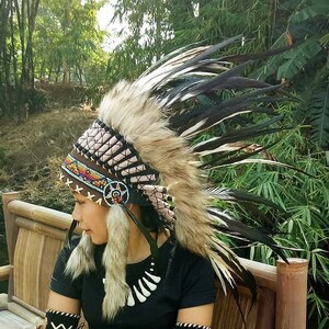 May include: A Native American headdress with brown, tan, and black feathers. The headdress features a beaded band with colorful accents and fur trim. The person is wearing a black shirt and matching armbands with decorative stitching.