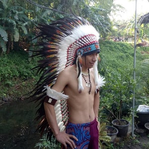 May include: A Native American headdress with white, black, and brown feathers. The headdress has a beaded band with red, blue, and white beads. The person is wearing a beaded necklace and armbands.