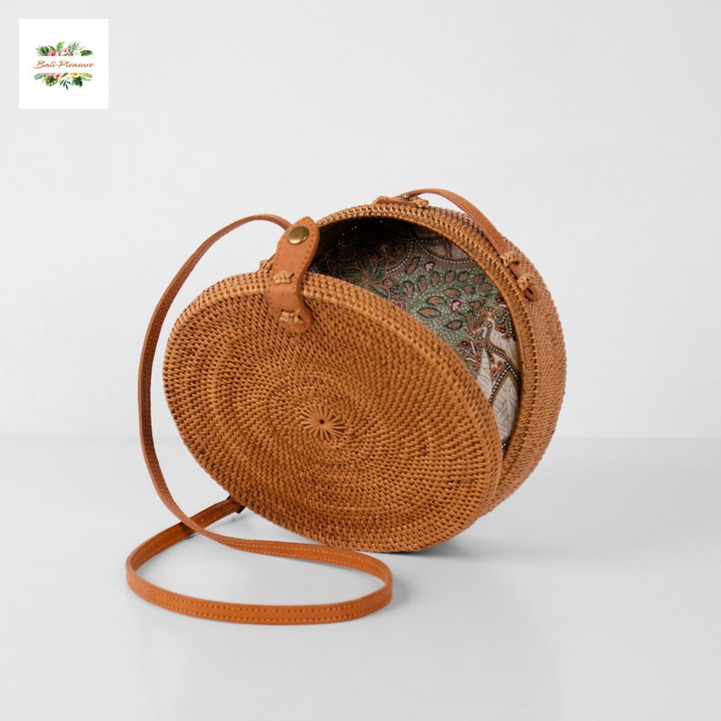 May include: A round, woven rattan bag with a leather strap and clasp. The bag's interior, visible when open, features a patterned fabric in shades of green, brown, and white. The bag is designed with a circular shape and a long, adjustable strap.