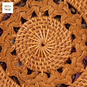 May include: Close-up of a round, woven rattan coaster with a central circular design and intricate woven patterns. The coaster is a warm, natural brown color, with a floral logo in the upper left corner. The coaster is approximately 10 cm in diameter.