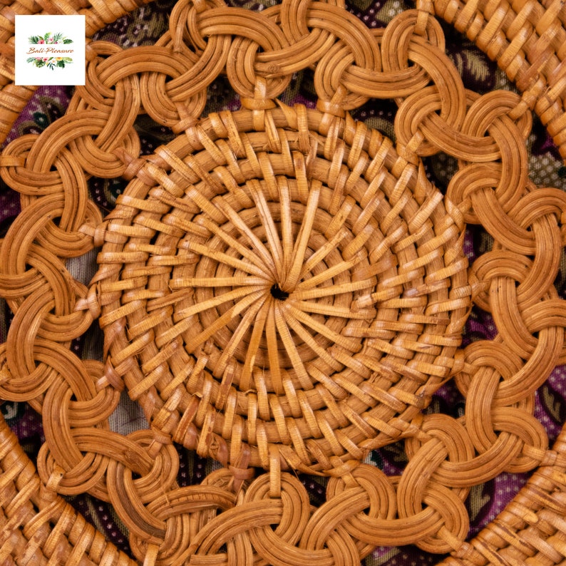 May include: Close-up of a round, woven rattan coaster with a central circular design and intricate woven patterns. The coaster is a warm, natural brown color, with a floral logo in the upper left corner. The coaster is approximately 10 cm in diameter.