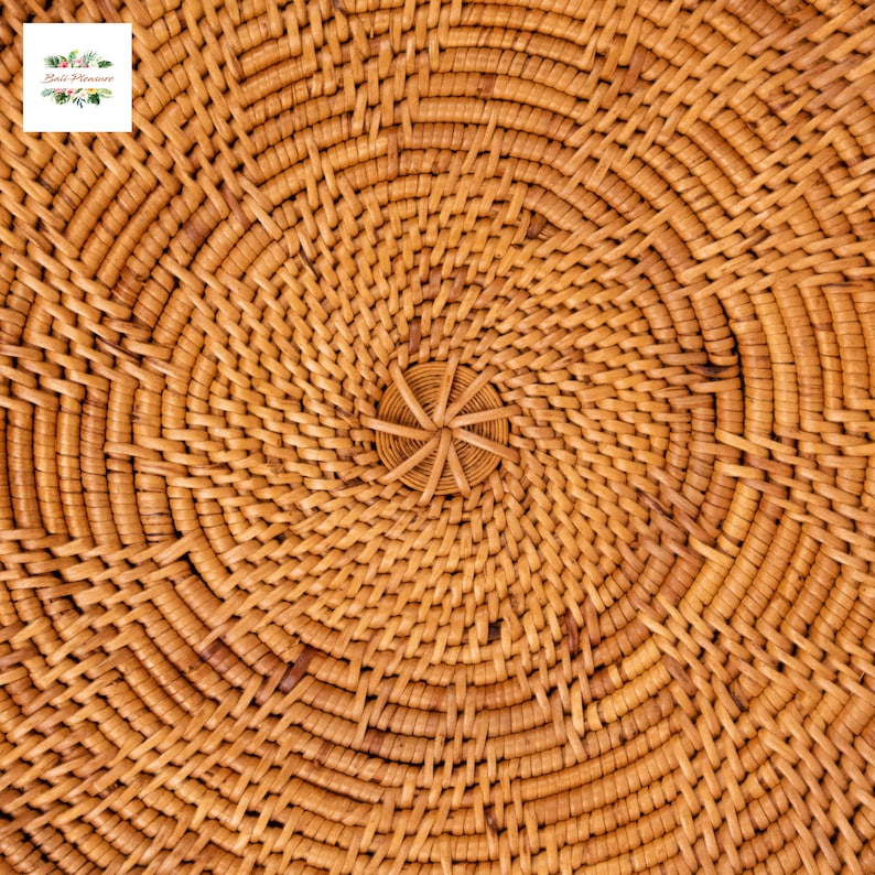Op de afbeelding: Close-up van een geweven rotan mand met een spiraalvormig ontwerp. Het natuurlijke bruine materiaal is strak geweven in concentrische cirkels, waardoor een textuur ontstaat. Het midden heeft een klein, stervormig detail. De mand is waarschijnlijk handgemaakt.
