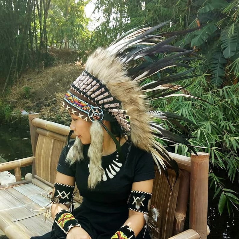 May include: A Native American headdress with brown, tan, and black feathers. The headdress features a beaded band with colorful accents and fur trim. The person is wearing a black shirt and matching armbands with decorative stitching.
