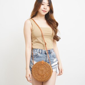 Op de afbeelding: Een ronde, geweven crossbodytas van rotan met een bruine leren riem. De tas heeft een natuurlijke bruine kleur en een cirkelvorm. De tas wordt over de schouder gedragen.