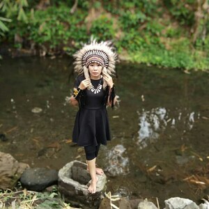 May include: A person wearing a Native American headdress with white and brown feathers, a black dress, and a white necklace. The person is standing in a body of water.