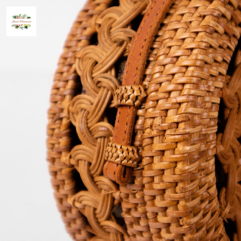 May include: Close-up of a round, woven rattan bag with a brown leather strap. The bag features intricate weaving patterns and a natural, light brown color. The strap is secured with decorative woven accents.