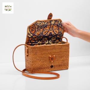 May include: A rectangular woven rattan handbag with a patterned interior lining. The bag has a leather strap and a leather clasp closure. The interior lining features a design in shades of blue, orange, and yellow. The bag is open, revealing the interior.