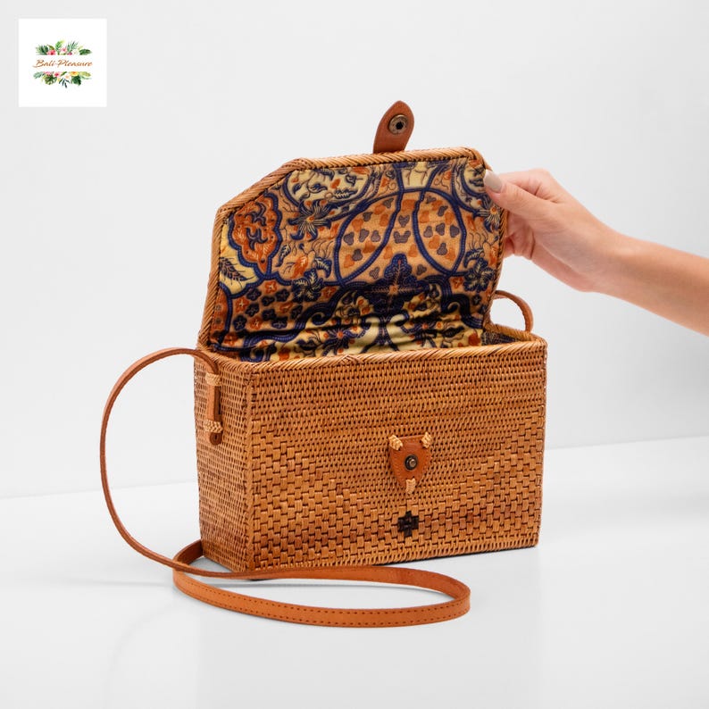 May include: A rectangular woven rattan handbag with a patterned interior lining. The bag has a leather strap and a leather clasp closure. The interior lining features a design in shades of blue, orange, and yellow. The bag is open, revealing the interior.