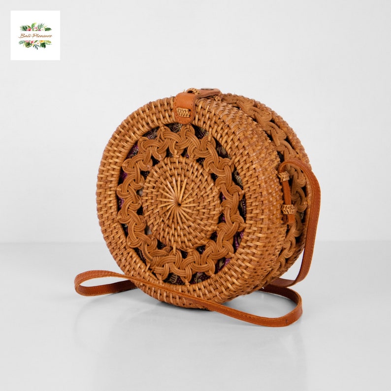 May include: A round, woven rattan handbag with a brown leather strap. The bag features a decorative, openwork design around the edges and a woven center. The bag is a natural tan color and has a leather closure.