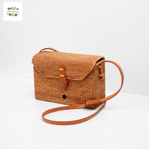 May include: Woven rattan crossbody bag in a rectangular shape with a flap closure. The bag features a brown leather strap and accents, including a button closure and a decorative emblem. The bag is a natural tan color.