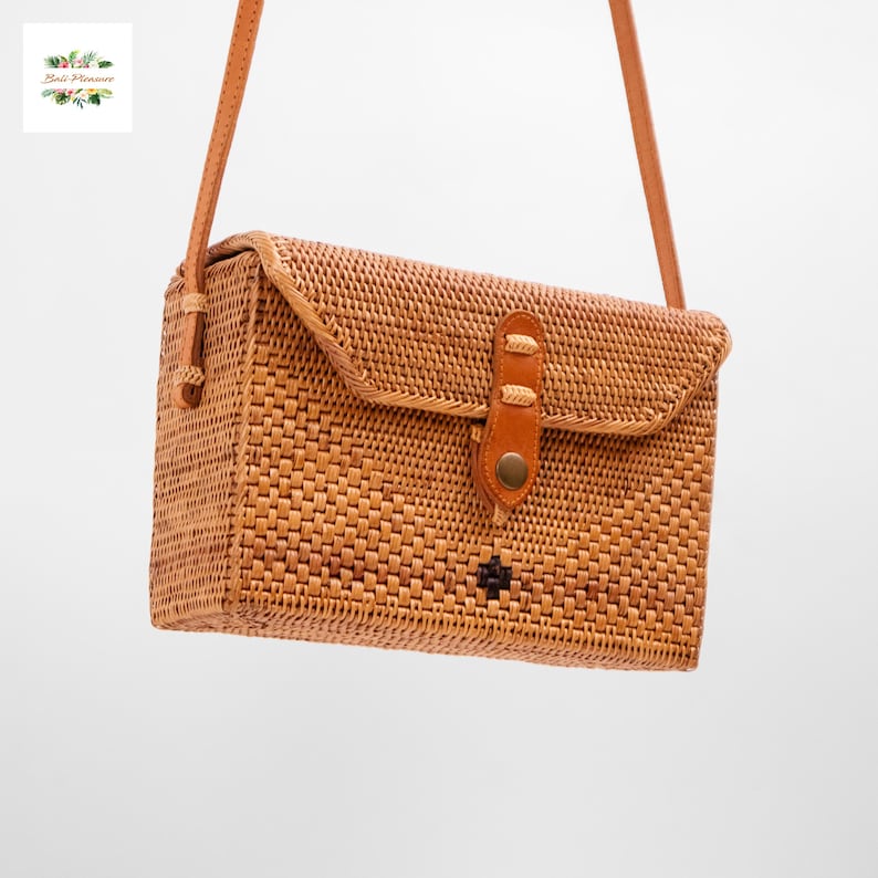 May include: A rectangular, woven rattan crossbody bag in a light brown hue. The bag features a flap closure secured with a leather strap and a button. A matching leather strap is attached for carrying. The bag has a small, dark-colored decorative element.