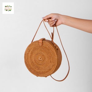 May include: A round, woven rattan bag with a brown leather strap and clasp. The bag is held by a hand against a white background. The bag has a spiral design in the center. The bag is a natural tan color.