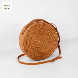 May include: A round, woven rattan bag with a brown leather strap and accents. The bag is tan in color and has a spiral weave pattern. The strap is adjustable with a buckle. The bag is designed to be worn over the shoulder.
