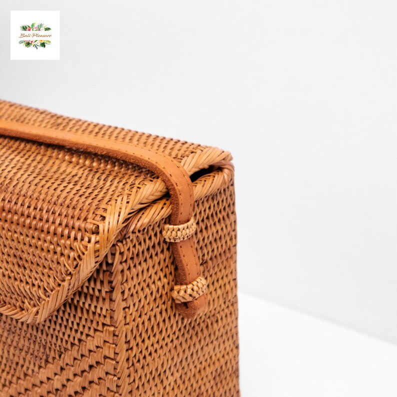 May include: A woven rattan handbag with a leather strap and clasp. The bag is a light brown color and has a rectangular shape. The strap is a lighter shade of brown. The bag is likely handmade and has a natural, rustic aesthetic.