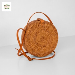 Op de afbeelding: Een ronde crossbody tas van geweven rotan in een natuurlijke bruine kleur. De tas heeft een bruine leren riem en een leren sluiting. De tas heeft een diameter van ongeveer 20 cm.