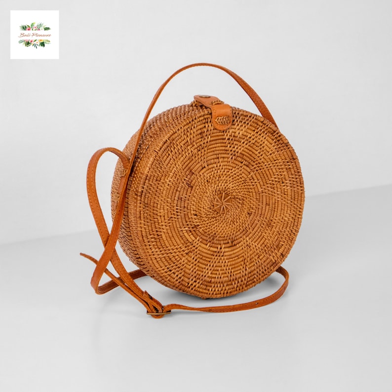 Op de afbeelding: Een ronde crossbody tas van geweven rotan in een natuurlijke bruine kleur. De tas heeft een bruine leren riem en een leren sluiting. De tas heeft een diameter van ongeveer 20 cm.
