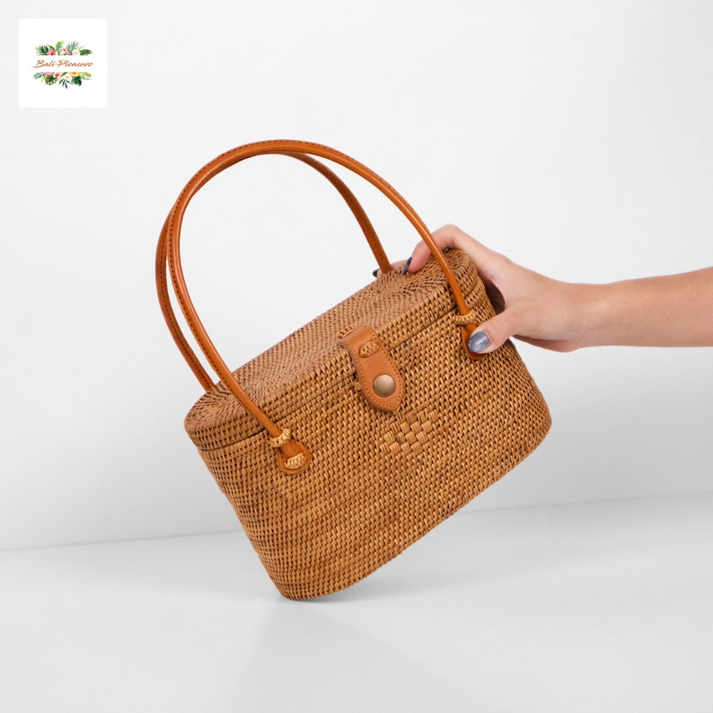 Puede incluir: Bolso de mano rectangular de rat&aacute;n tejido, con asa de cuero marr&oacute;n y cierre de cuero. El bolso es de color marr&oacute;n claro y tiene un patr&oacute;n tejido. El asa es arqueada y se sujeta al bolso con peque&ntilde;as trabillas de cuero.