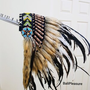 May include: A Native American headdress with a black beaded headband and a colorful beaded medallion. The headdress features brown, white, and black feathers, and a fur accent. The text "BaliPleasure" is visible.