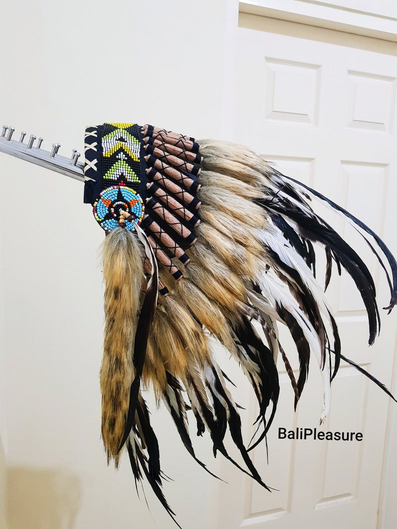 May include: A Native American headdress with a black beaded headband and a colorful beaded medallion. The headdress features brown, white, and black feathers, and a fur accent. The text "BaliPleasure" is visible.