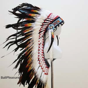 Op de afbeelding: Een Indiaanse hoofdtooi met witte, rode, zwarte en gele veren. De hoofdtooi wordt getoond op een witte mannequinhoofd. De tekst "BaliPleasure" is zichtbaar linksonder in de afbeelding.