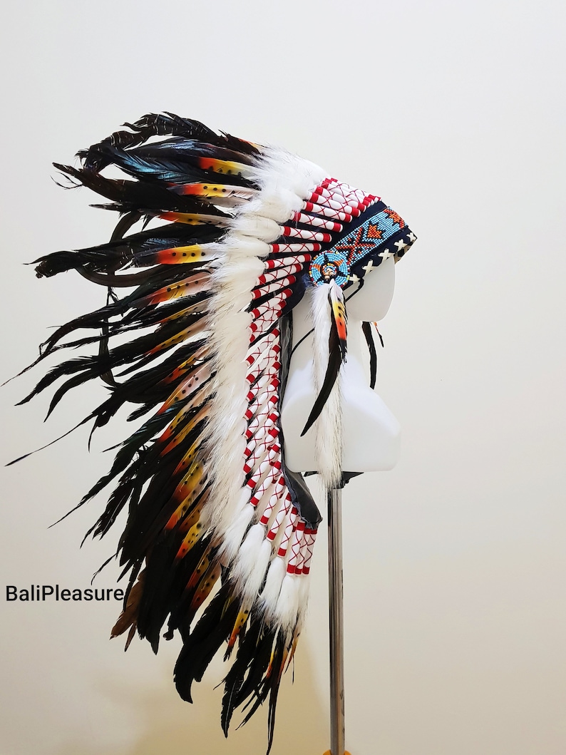 Op de afbeelding: Een Indiaanse hoofdtooi met witte, rode, zwarte en gele veren. De hoofdtooi wordt getoond op een witte mannequinhoofd. De tekst "BaliPleasure" is zichtbaar linksonder in de afbeelding.