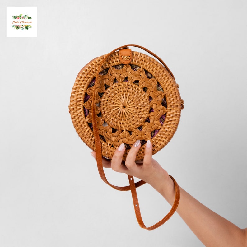 May include: A round, woven rattan bag with a brown leather strap. The bag features a detailed, circular pattern in the center and a decorative border. The bag is held up by a person's hand. The bag is approximately 8 inches in diameter.