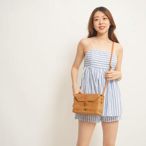 May include: A light blue and white striped camisole top and shorts set. A woven, rectangular crossbody bag with a leather strap and a flap closure is worn over the shoulder. The bag is made of natural-colored woven material.