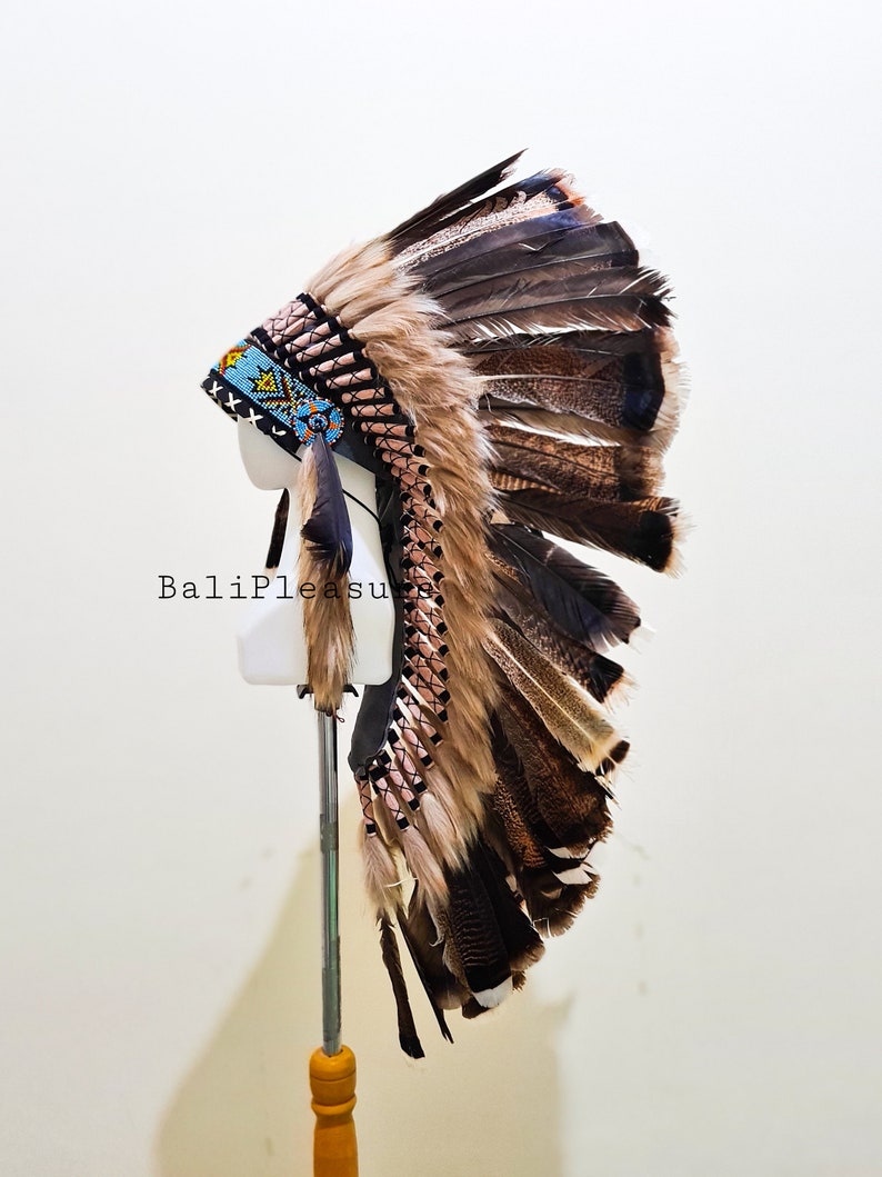 May include: A Native American headdress with brown and black feathers, displayed on a mannequin head. The headdress features a beaded band with blue, yellow, and black accents. The text "BaliPleasure" is visible on the image.