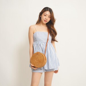 May include: A light blue and white striped romper with spaghetti straps and matching shorts. A round, woven rattan crossbody bag with a brown leather strap is worn over the shoulder. The outfit is casual and summery.
