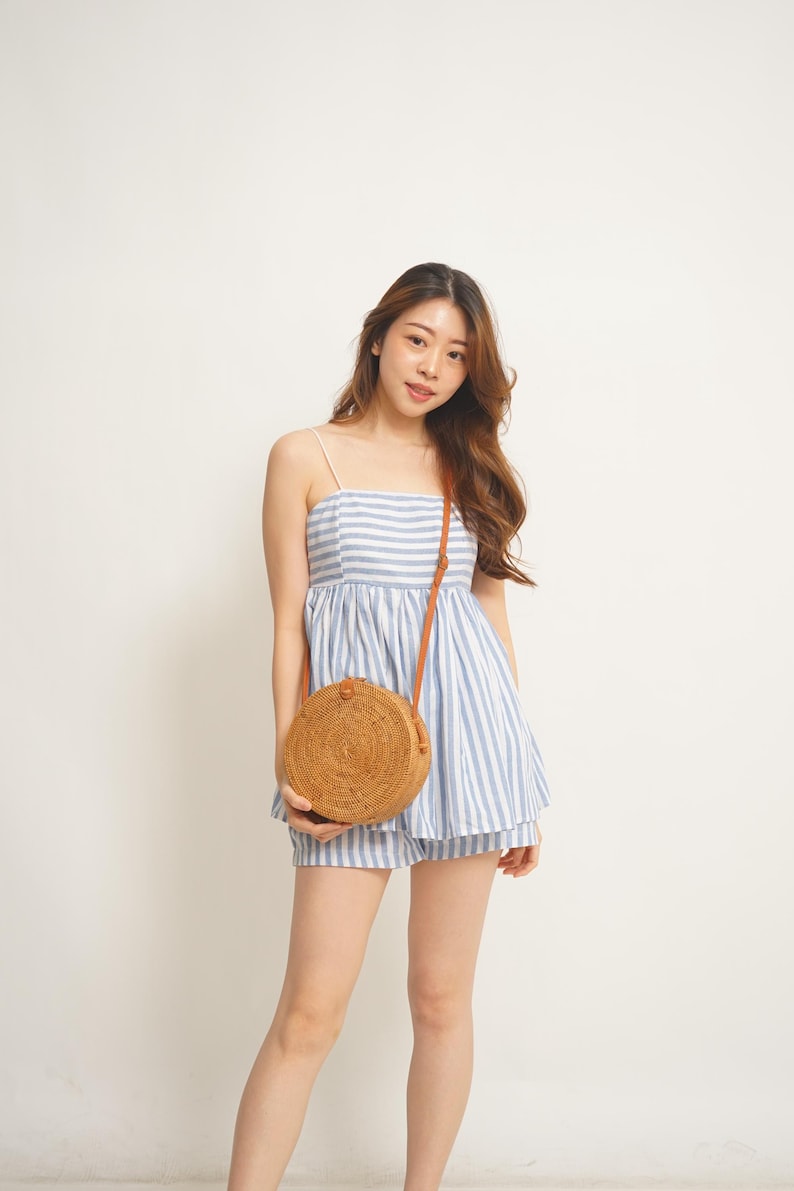 May include: A light blue and white striped romper with spaghetti straps and matching shorts. A round, woven rattan crossbody bag with a brown leather strap is worn over the shoulder. The outfit is casual and summery.
