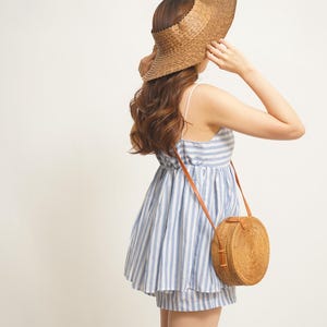 May include: A woman wearing a blue and white striped dress and shorts, a woven sun visor, and a round, woven crossbody bag with a brown leather strap. The dress has thin straps and a gathered waist.