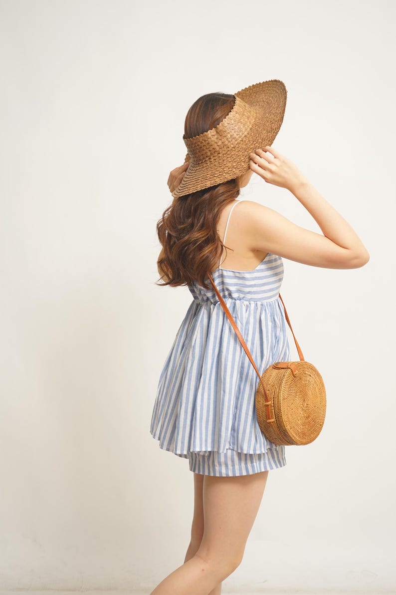 May include: A woman wearing a blue and white striped dress and shorts, a woven sun visor, and a round, woven crossbody bag with a brown leather strap. The dress has thin straps and a gathered waist.